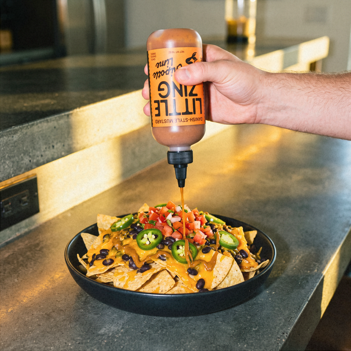 A hand squeezes a bottle of Little Zing Hot Sauce onto a loaded plate of nachos on a dark countertop in a kitchen setting.