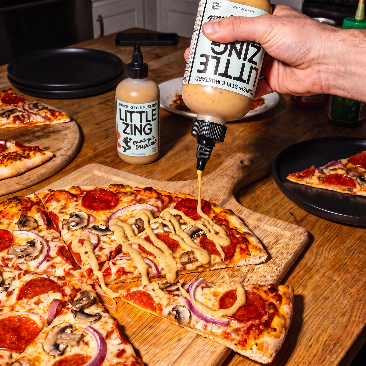 A hand holds a squeeze bottle of Little Zing Originals garlic aioli dressing, drizzling it over a slice of pepperoni and mushroom pizza on a wooden board. Another bottle of the aioli sits on the table, with other pizza slices and black plates around it on a wooden countertop.